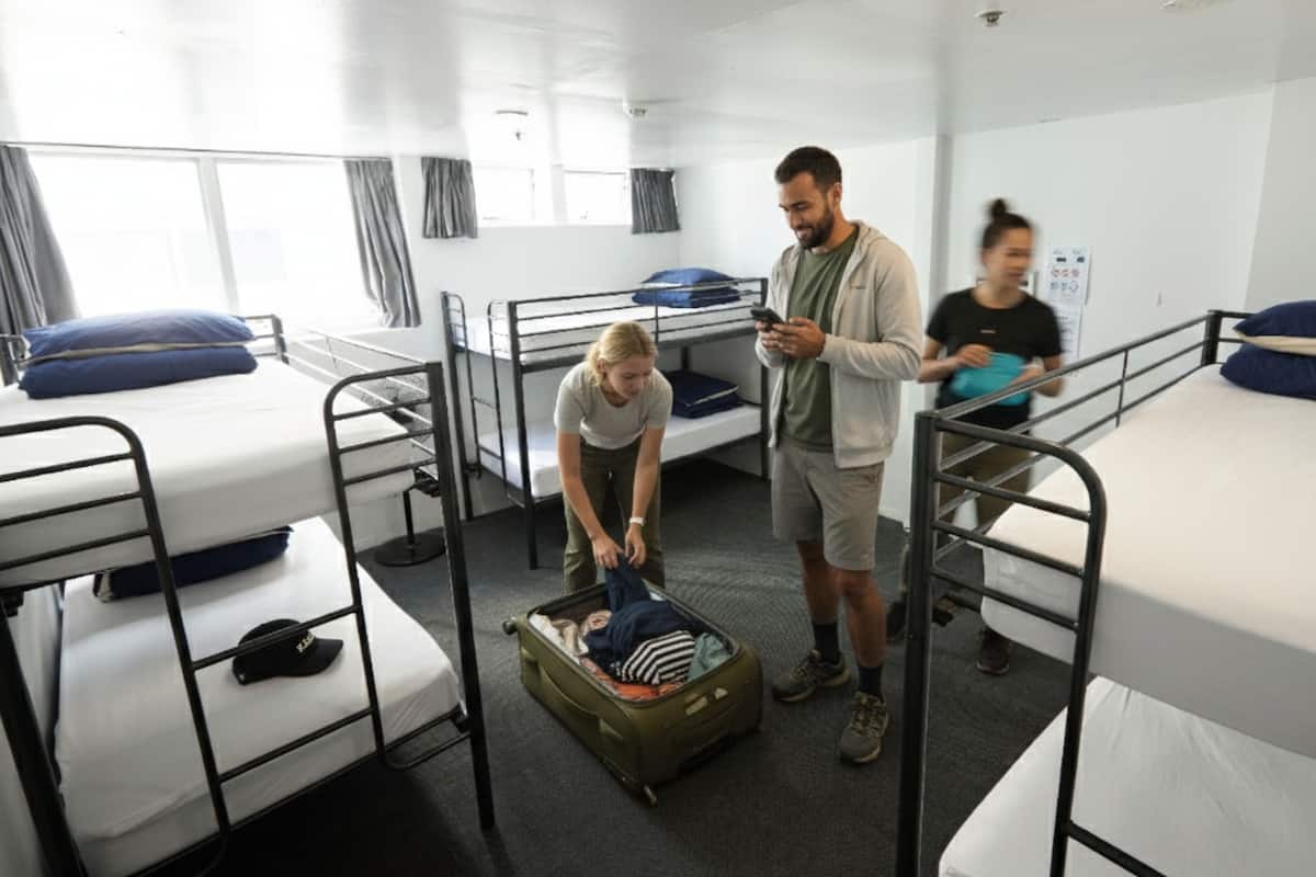 Une chambre de dortoir avec des lits superposés, une valise posée sur le sol et une personne debout près du lit.
