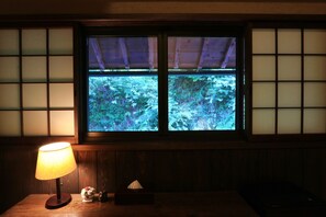 A room with a wooden table, a lamp, and a window with a view of greenery.