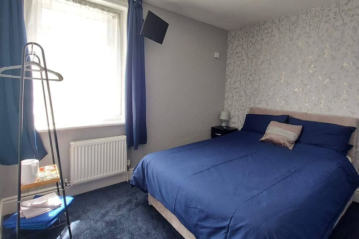 Un dormitorio con cama, una silla colgante, radiador, televisión y ventana con cortinas.