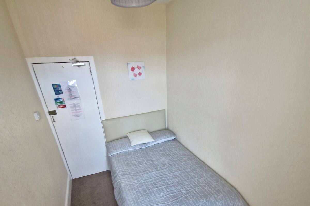 Una habitación pequeña con cama individual, colcha de cama gris con estampado, puerta blanca con un tablón de anuncios y una lámpara colgante gris.