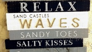 Une pancarte avec des inscriptions telles que « PLAGE », « SOLEIL », « DÉTENTE », « VAGUES », « PIEDS DANS LE SABLE », « BAISERS SALÉS » et « PARADIS ».
