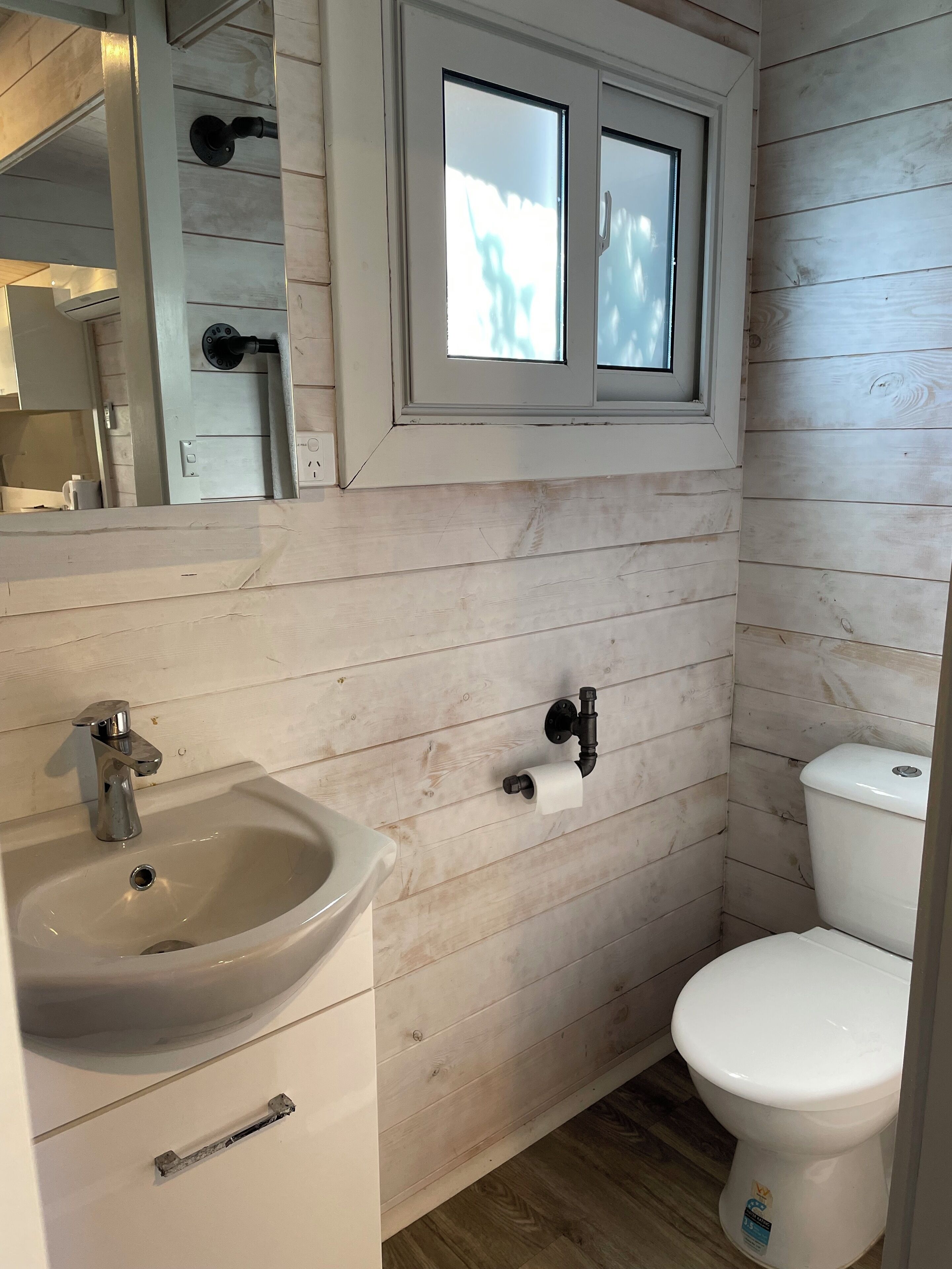 A bathroom with a wooden wall, a sink, a toilet, and a window above the toilet.