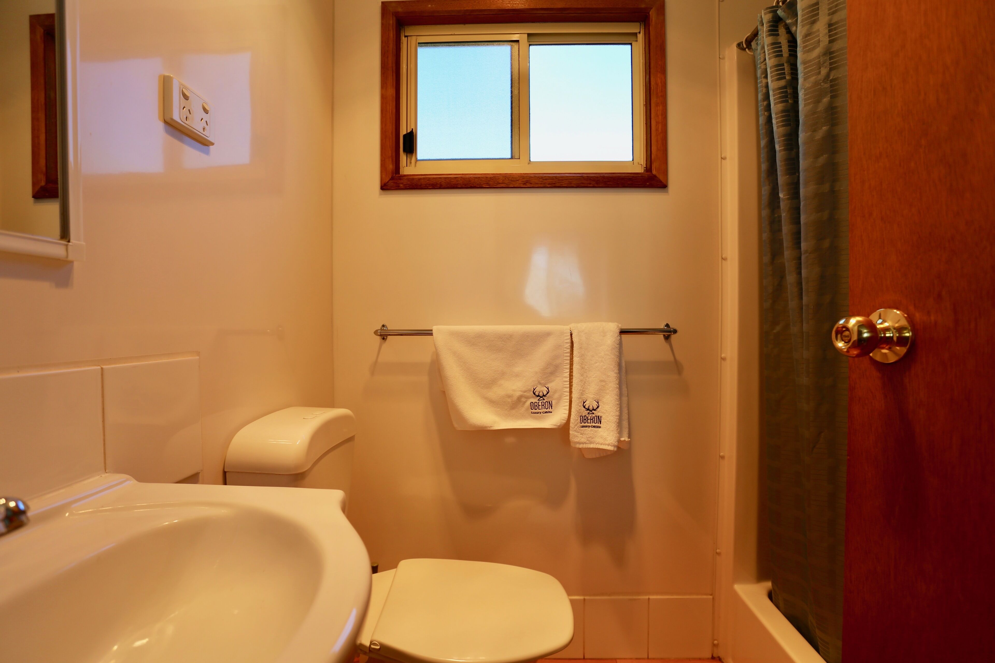 Casa de banho com lavatório branco, sanita e banheira. Há uma janela sobre a banheira e uma toalha pendurada num porta-toalhas.