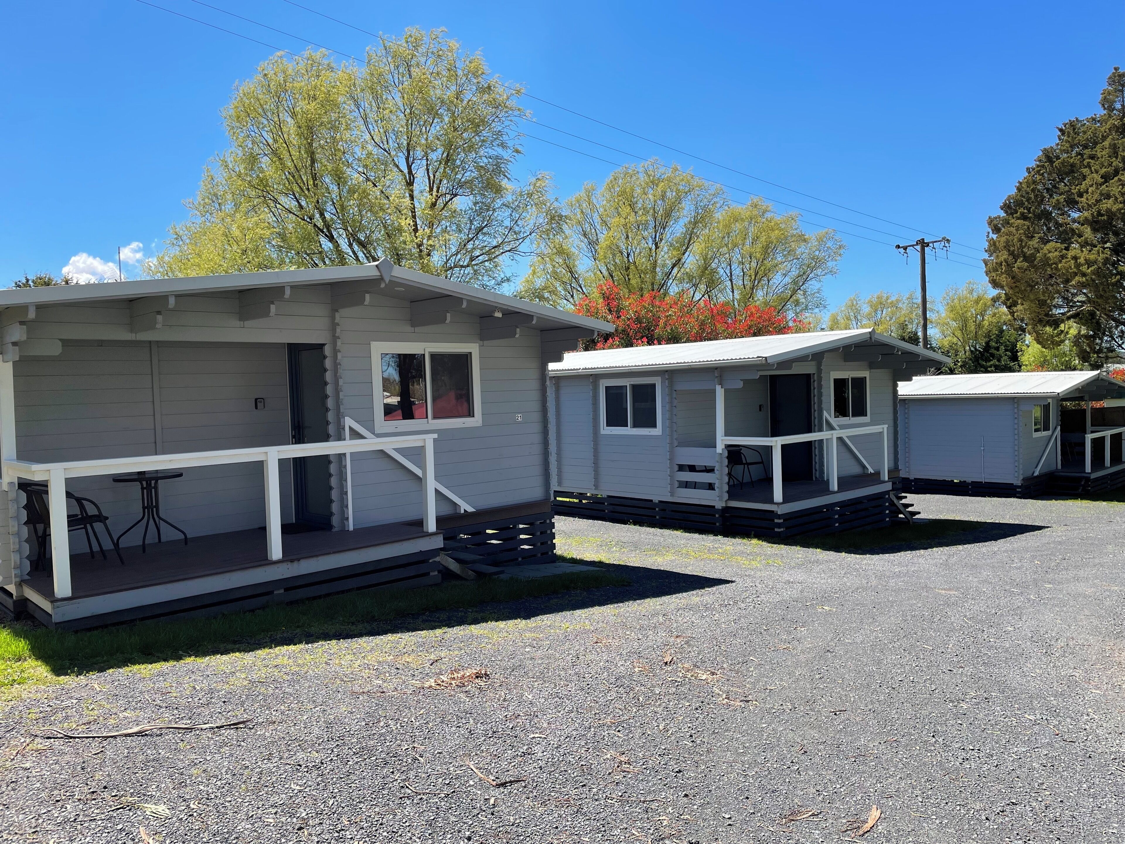 A row of small, single-story cabins with a porch and a table, set in a gravel area with trees in the background.