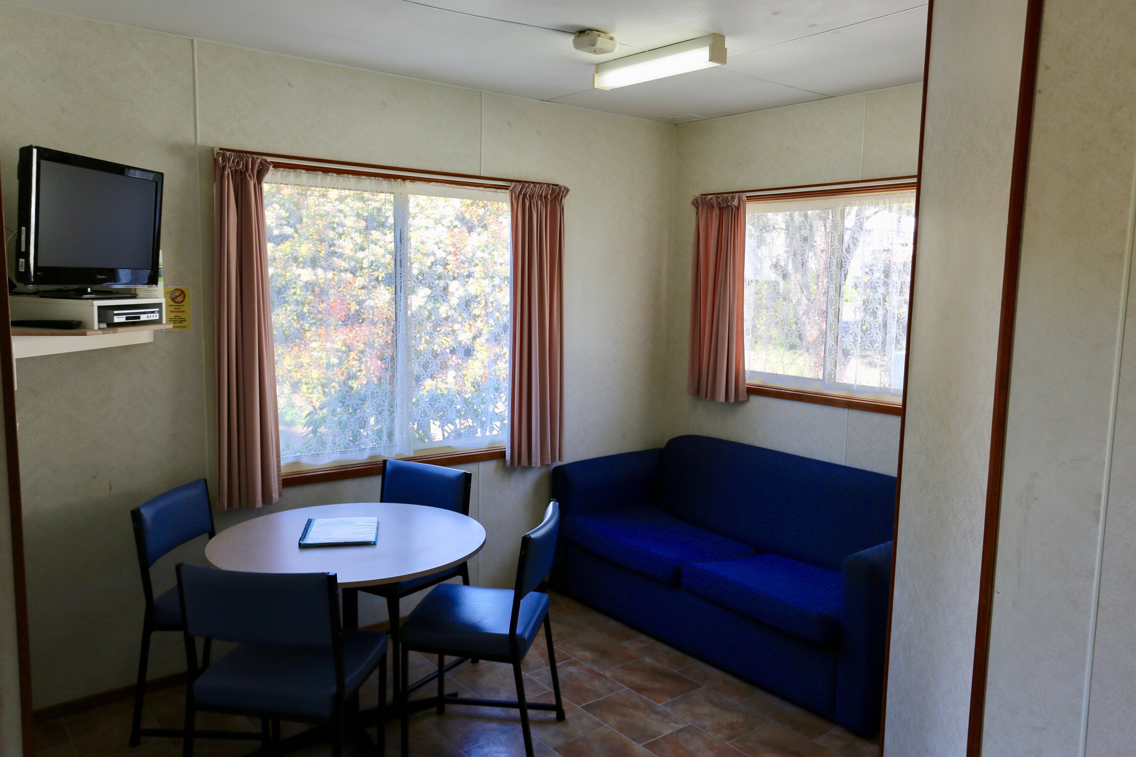 A room with a blue sofa, a dining table with chairs, a television on a shelf, and windows with curtains.