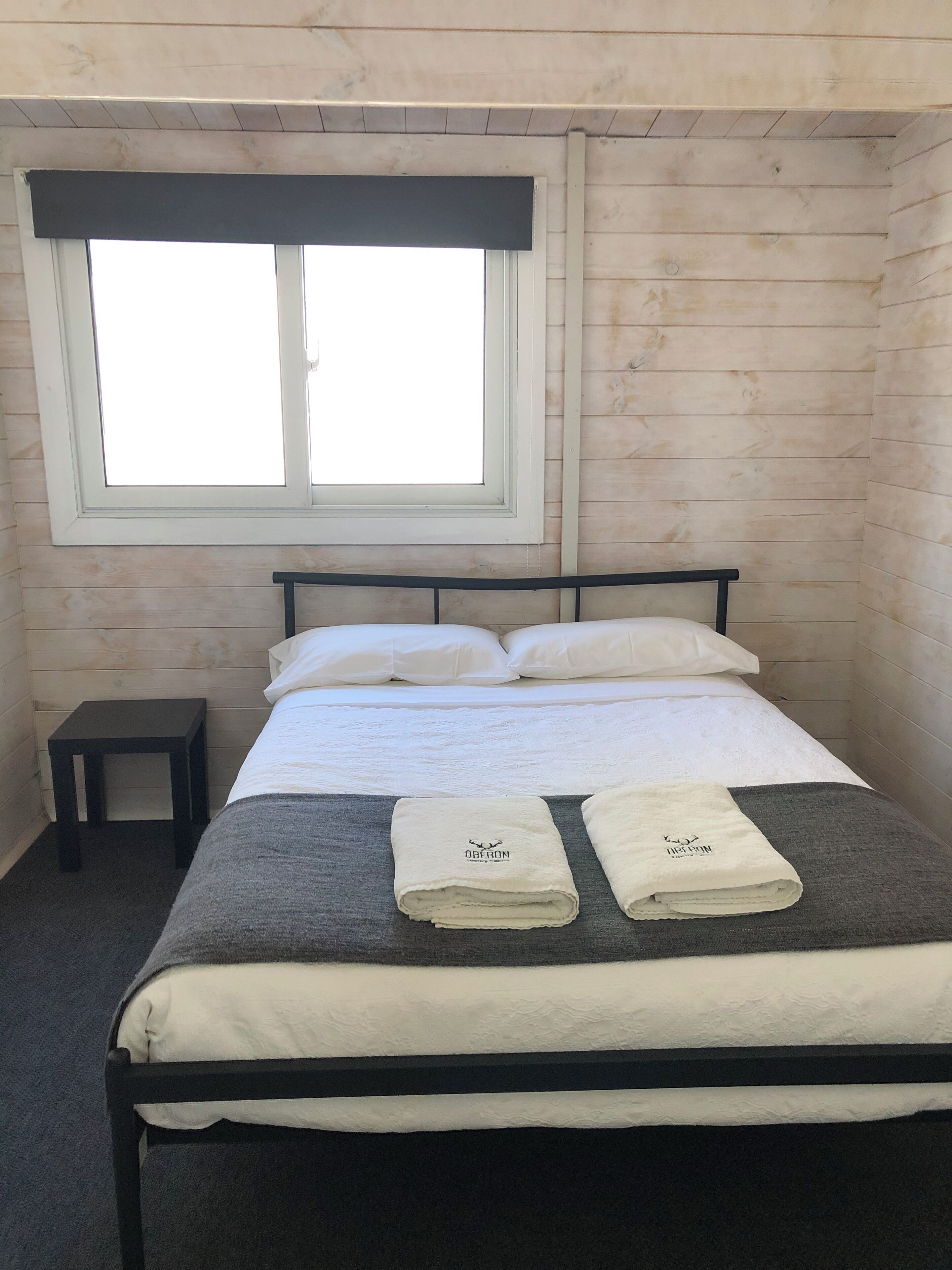 A neatly made bed with two towels, a small bedside table, and a window with blinds.