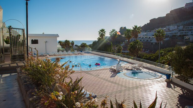 A swimming pool with two sections, surrounded by lounge chairs and plants, with a view of the sea and buildings in the background. - Apartamanto with terrace, free WiFi, communal pool in Puerto Rico (Mogán)