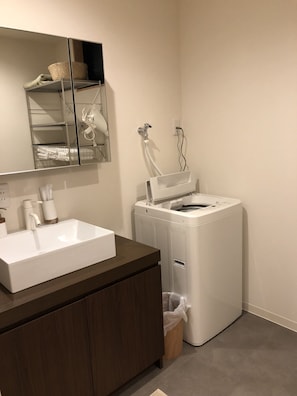 A modern bathroom with a white sink, a mirror, a built-in washing machine, and a wooden vanity.