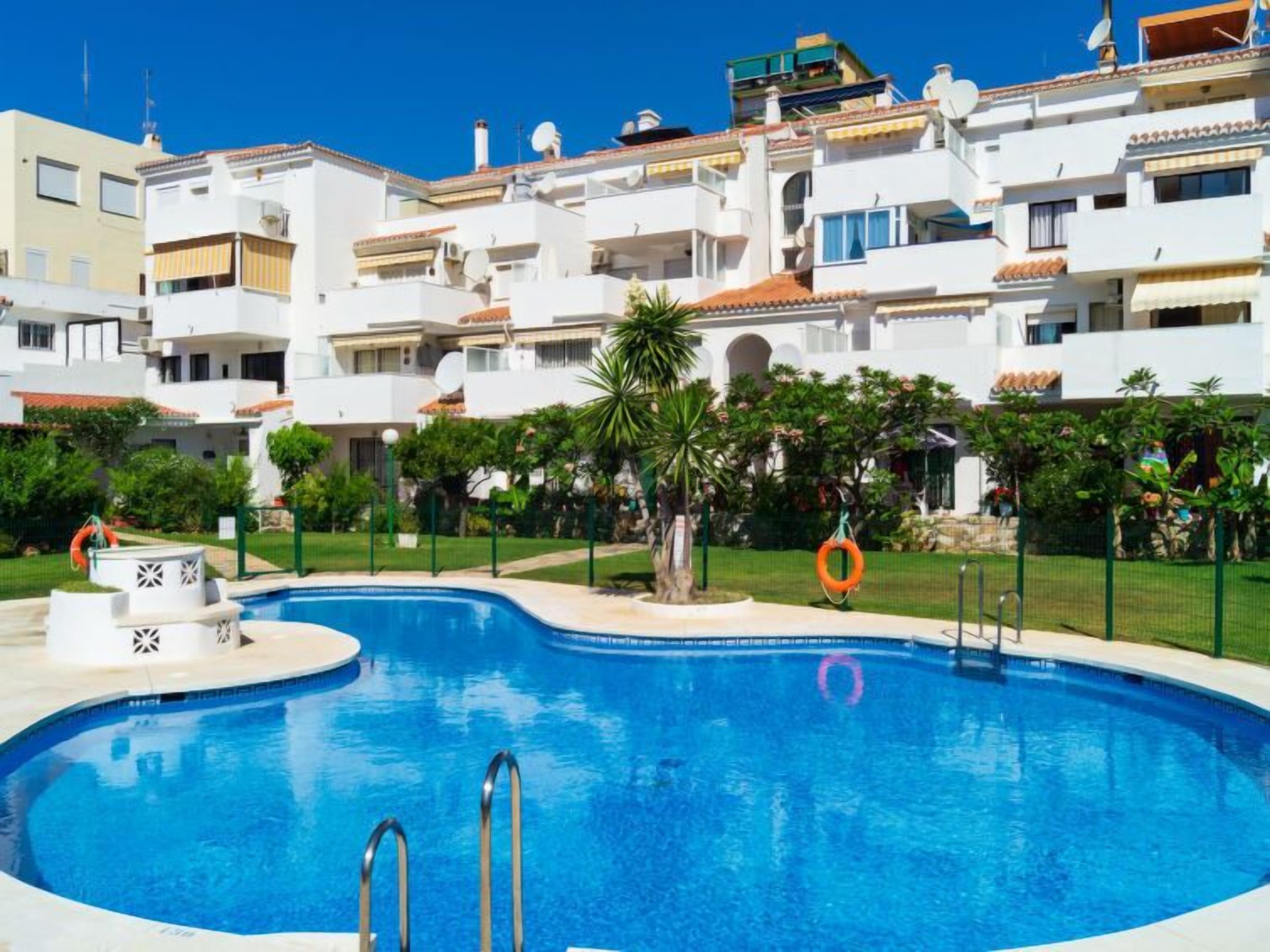 A swimming pool surrounded by a white building with balconies and a clear blue sky.