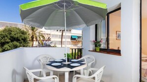 En balkon med et bord og stole, en grøn og hvid parasol, samt et vindue der ud til bygninger og træer.