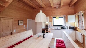 A wooden cabin interior with a dining area, kitchen, and a window with curtains.