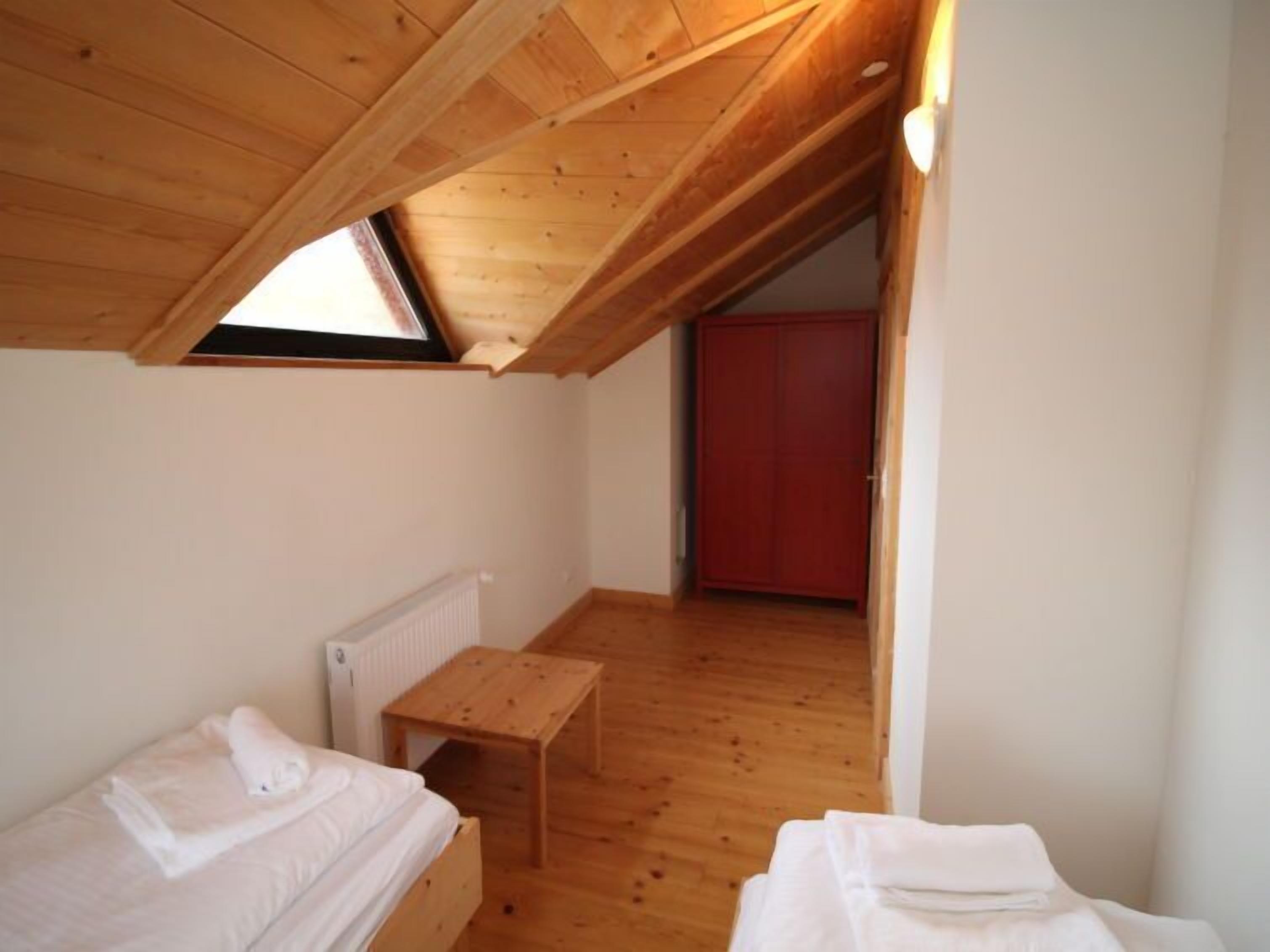 Une petite cabane d’une seule pièce, avec un plafond en pente, un plancher en bois, une petite table et un lit près d’une fenêtre.