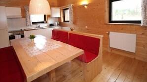 A wooden cabin interior with a kitchen, a dining area with a bench, and a window providing natural light.