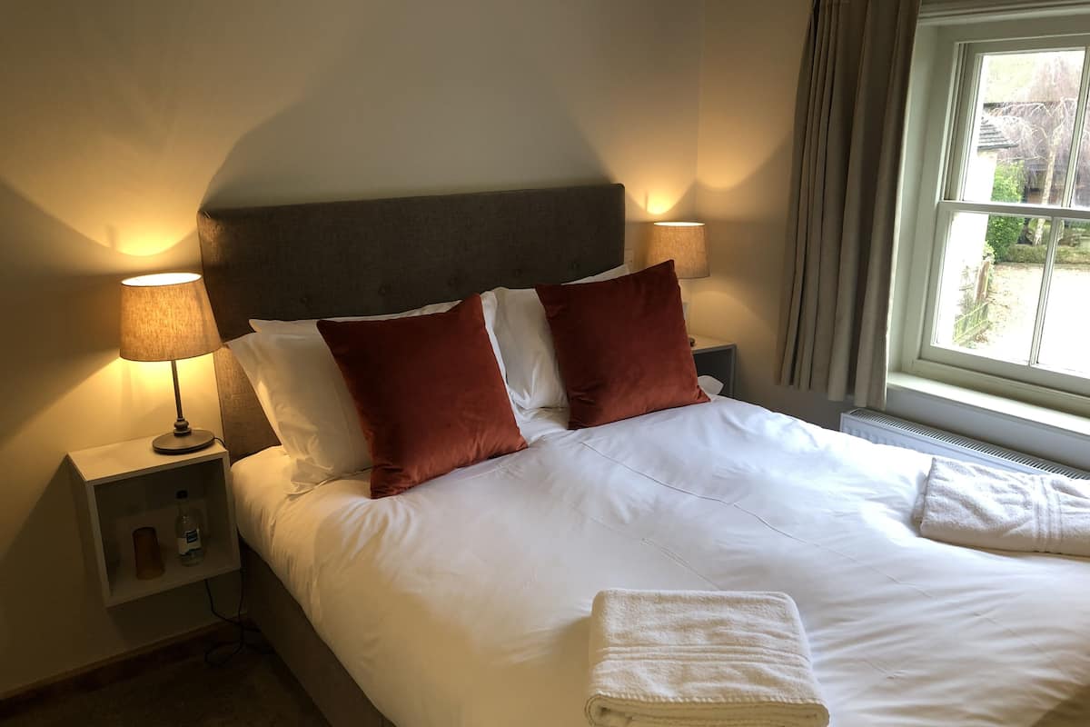 Ein ordentlich bezogenes Bett mit weißen Leinen und zwei braunen Kissen, eine Nachttischlampe und ein Fenster mit Vorhängen.