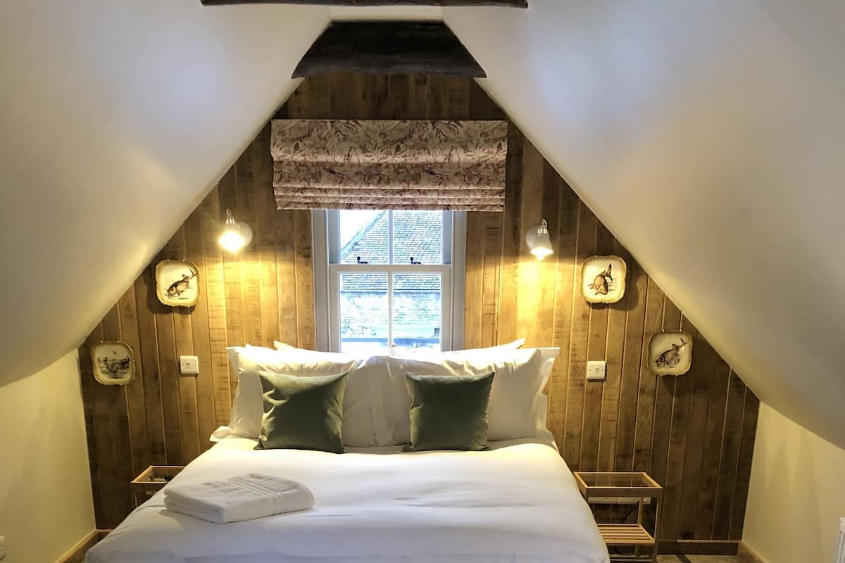 Ein Schlafzimmer mit schrägen Wänden, einem Bett mit weißer Bettwäsche, zwei grünen Kissen und einem Fenster mit Blick.