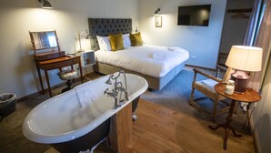 A hotel room with a large bathtub, two beds, a wooden floor, a desk with a mirror, and a TV on the wall. - Black Horse (Arundel)