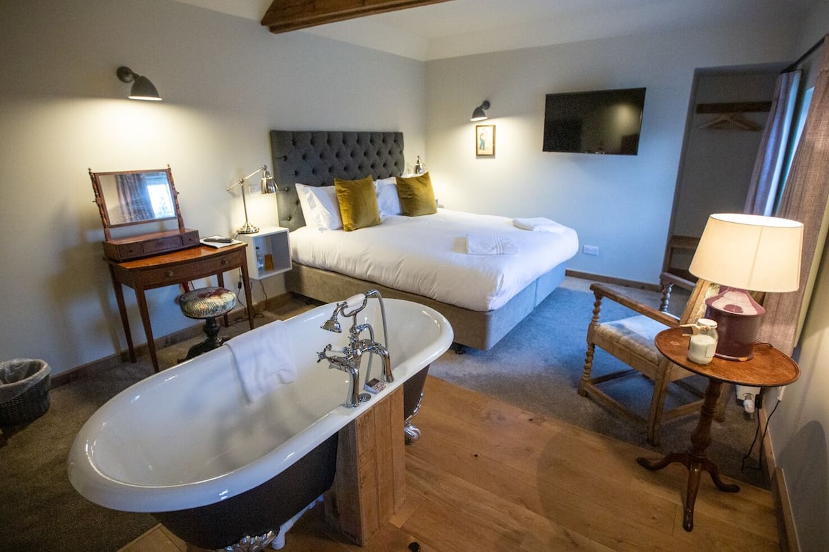 Ein Hotelzimmer mit einer großen Badewanne, zwei Betten, Holzboden, einem Schreibtisch mit Spiegel und einem an der Wand montierten Fernseher.