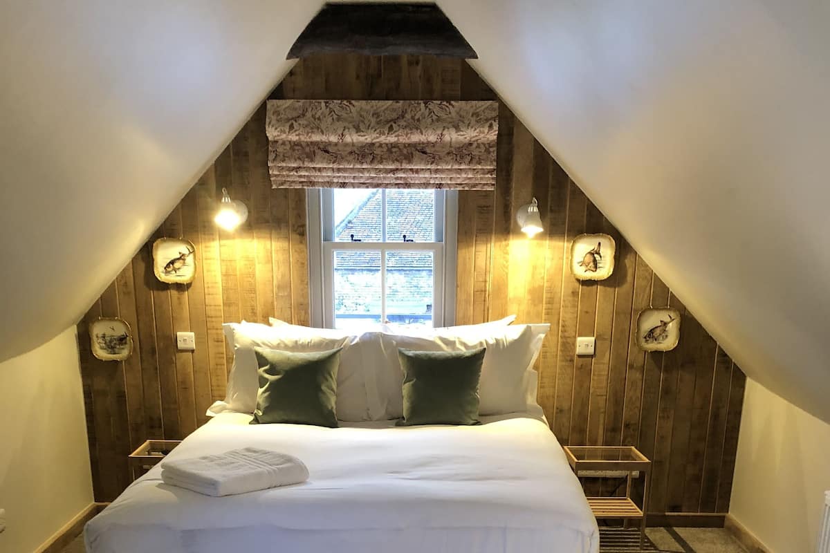 Ein Schlafzimmer mit schrägemausertem Dach, einem großen Bett, zwei Nachttischlampen und einem Fenster mit Blick.