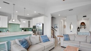 Una cocina moderna con armarios blancos, una isla central y electrodomésticos de acero inoxidable. En la zona de estar, hay un sofá color beige con cojines azules y una mesa de centro de madera.