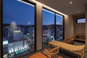 Habitación de hotel moderna con ventanas amplias que ofrecen vista a la ciudad, una pequeña mesa, sillas de mimbre y un televisor.