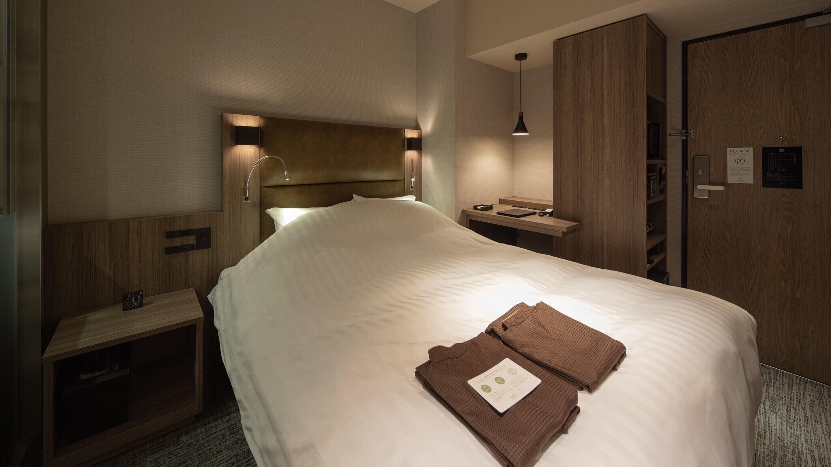 A hotel room with a large bed, a bedside table, a nightstand, a wardrobe, and a desk with a lamp.
