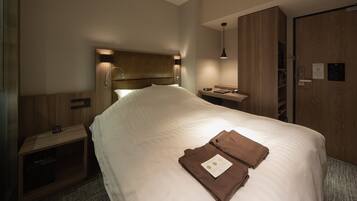 A hotel room with a large bed, a bedside table, a nightstand, a wardrobe, and a desk with a lamp.