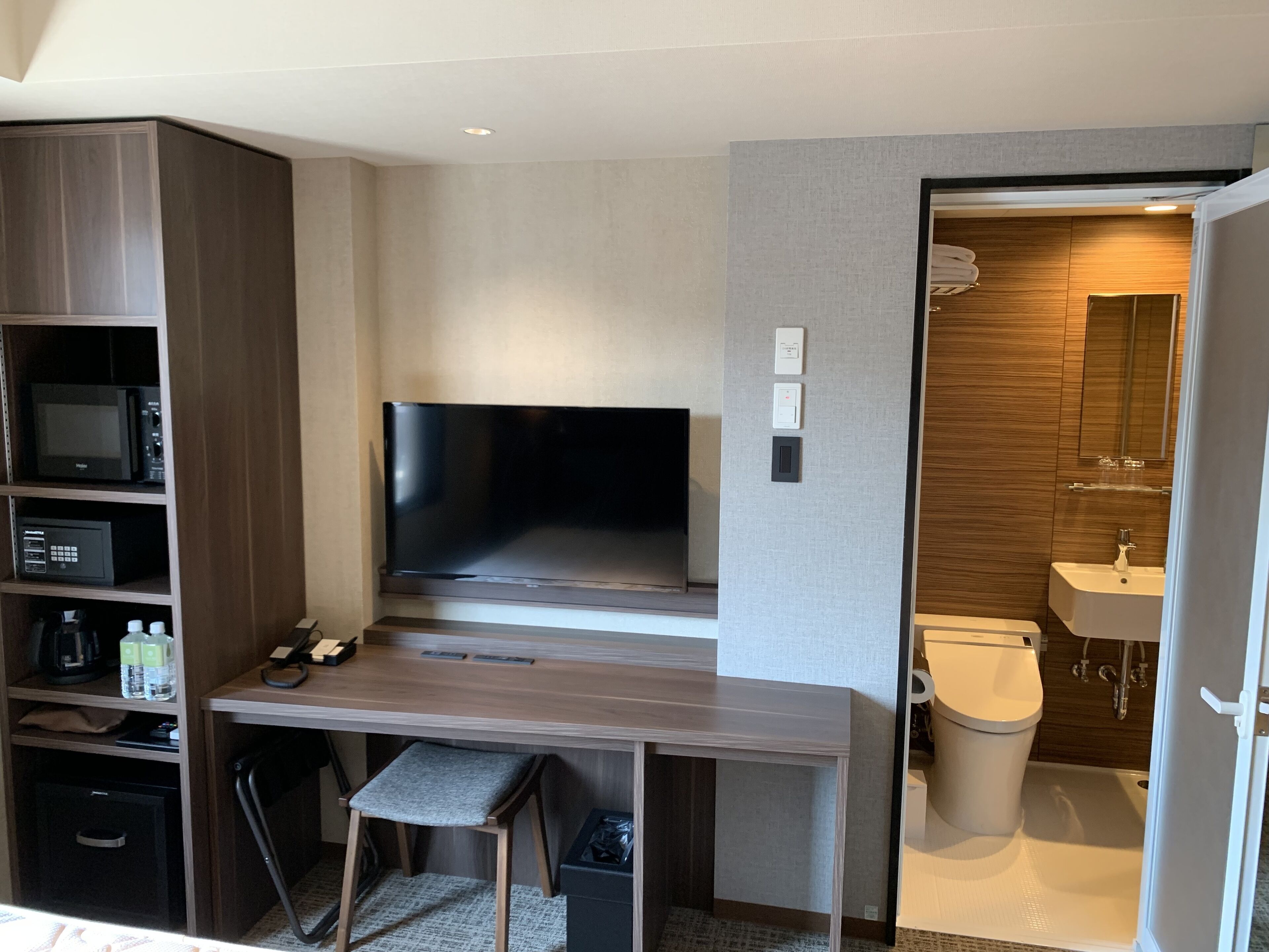 A modern hotel room with a flat-screen TV, built-in shelving, a desk, a chair, a trash bin, and a bathroom with a toilet, sink, and mirror.
