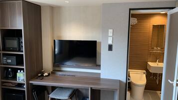 A modern hotel room with a flat-screen TV, built-in shelving, a desk, a chair, a trash bin, and a bathroom with a toilet, sink, and mirror.