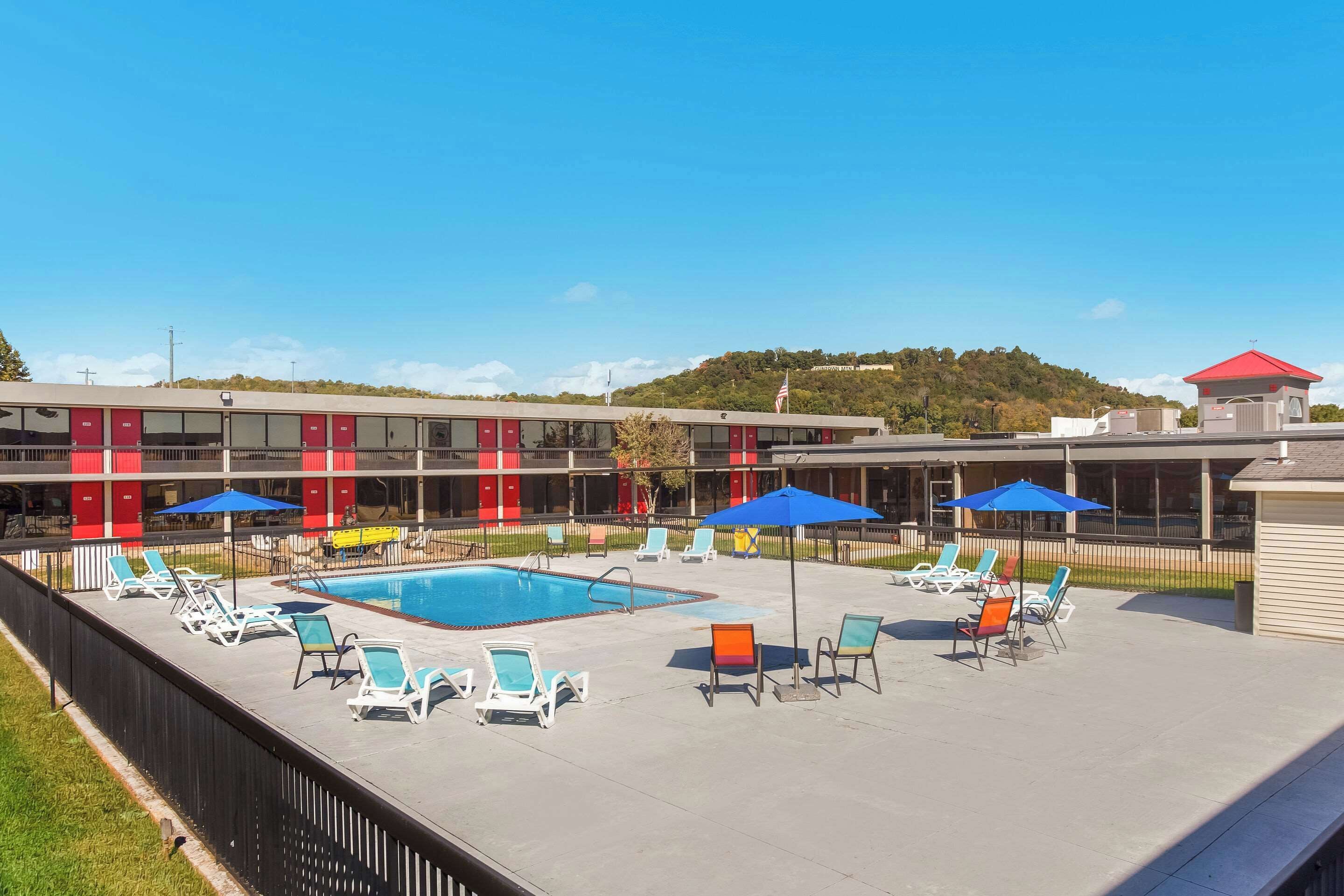 A pool area with lounge chairs and umbrellas, a building with red and beige exterior, and a flag on a hill in the background.