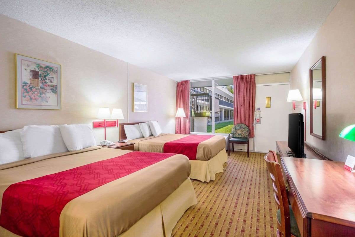 Ett hotellrum med två sängar, ett skrivbord, en stol, en TV och ett fönster med gardiner.