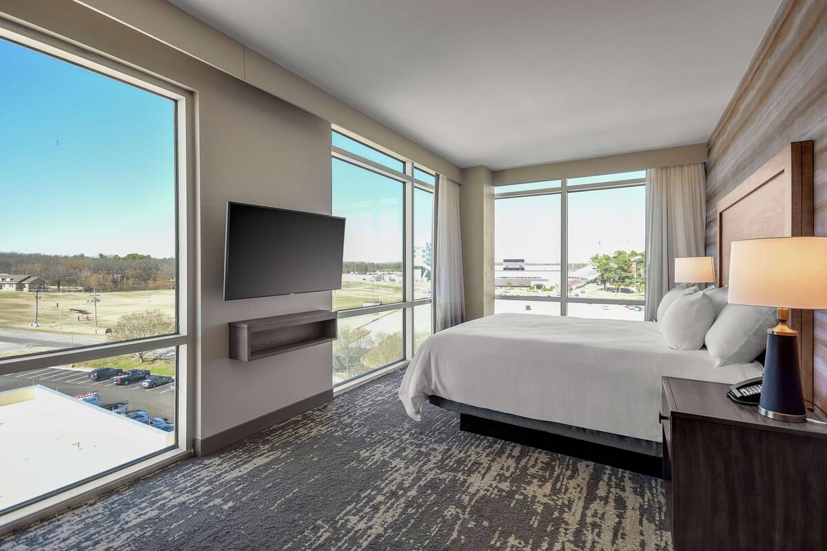 Hotelkamer met een groot bed, een flatscreen televisie aan de muur, een nachtkastje met een lamp, en uitzicht op een parkeerplaats en gebouwen door grote ramen.