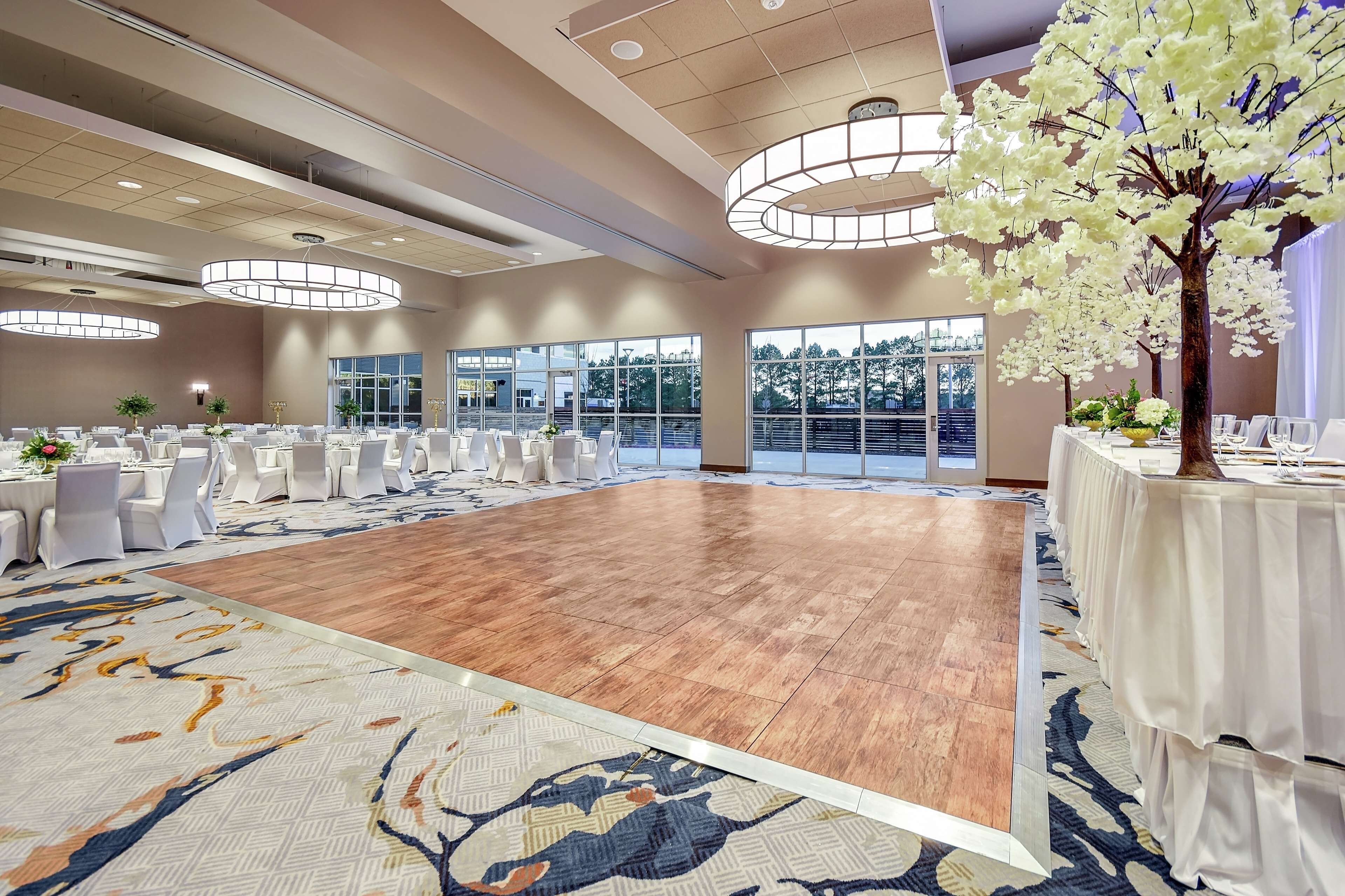 A spacious banquet hall with a wooden floor, round tables covered with white tablecloths, and large windows offering a view of the outdoors.