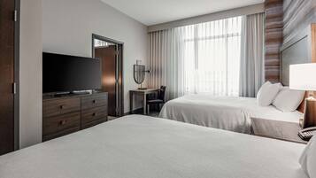 A hotel room with two beds, a TV, a desk with a chair, a mirror, and a large window with curtains.