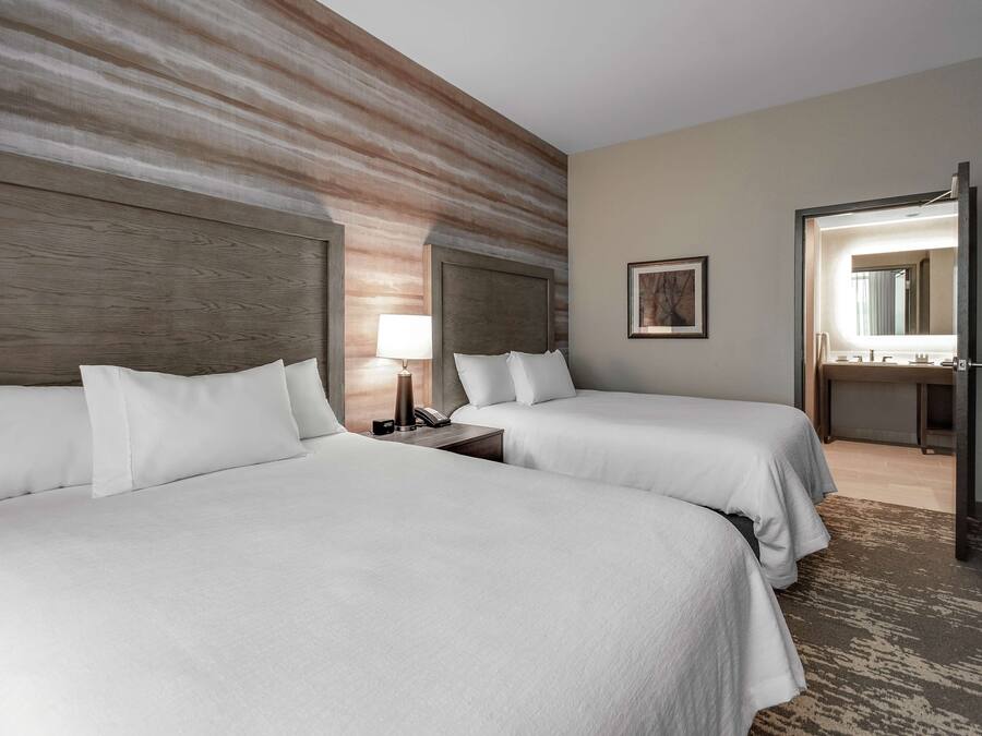 Habitación de hotel con dos camas, una mesita de noche con lámpara, un cuadro en la pared y un baño visible a través de una puerta abierta.