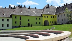 Un patio con un área de estar exterior moderna, rodeado de edificios de varios pisos con fachada verde y techos grises.