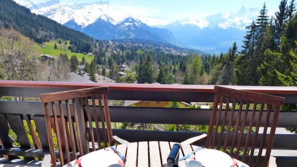 Una terraza de madera con dos sillas y una mesa para dos, con vistas a un paisaje montañoso de picos nevados y un valle abajo.