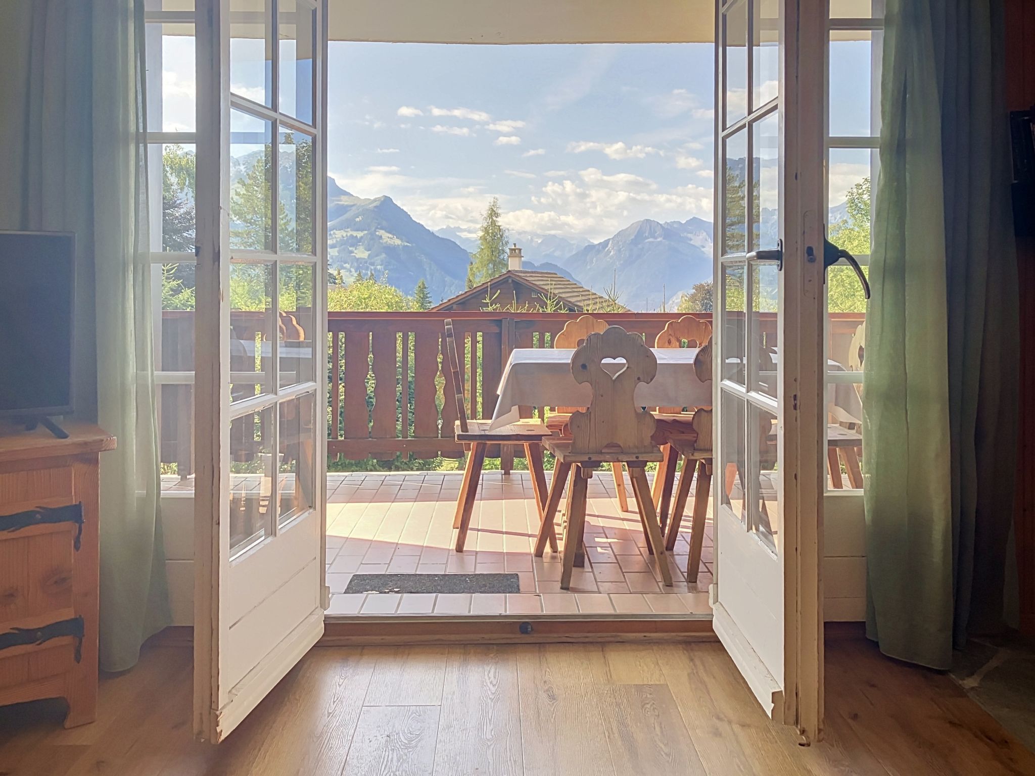 An open door leading to a balcony with a wooden table and chairs, a view of mountains, and a wooden deck.