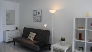 Una sala moderna con un sofá gris, una mesita auxiliar blanca con una planta, una lámpara de pie y una estantería blanca con libros y adornos.
