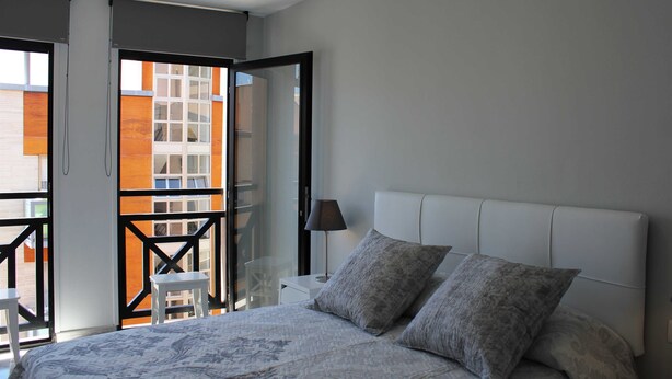 Un dormitorio con una cama grande, dos almohadas, una mesita de noche, una lámpara y vistas de un edificio a través de una puerta abierta.
