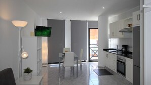 Una cocina moderna con armarios blancos, una zona de comedor con mesa y sillas, un televisor en la pared y una lámpara de pie.