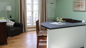 Un saló amb un sofà, una taula de dinar i una cadira. Hi ha una finestra amb cortines, una làmpada i un quadre al mur.