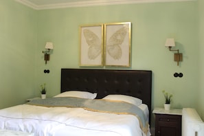 Cama bem arrumada com lençóis brancos e manta cinza e dourada. Existem duas luminárias fixadas na parede em ambos os lados da cama, e um par de quadros com borboletas acima dela.