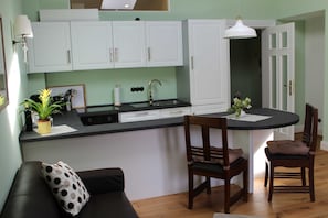 Uma cozinha compacta com armários brancos, bancada preta e um pequeno espaço de refeições com duas cadeiras de madeira.