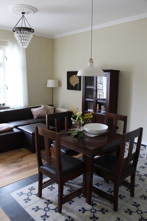 Een eetgedeelte met een houten tafel en stoelen, een kroonluchter en een boekenkast op de achtergrond.