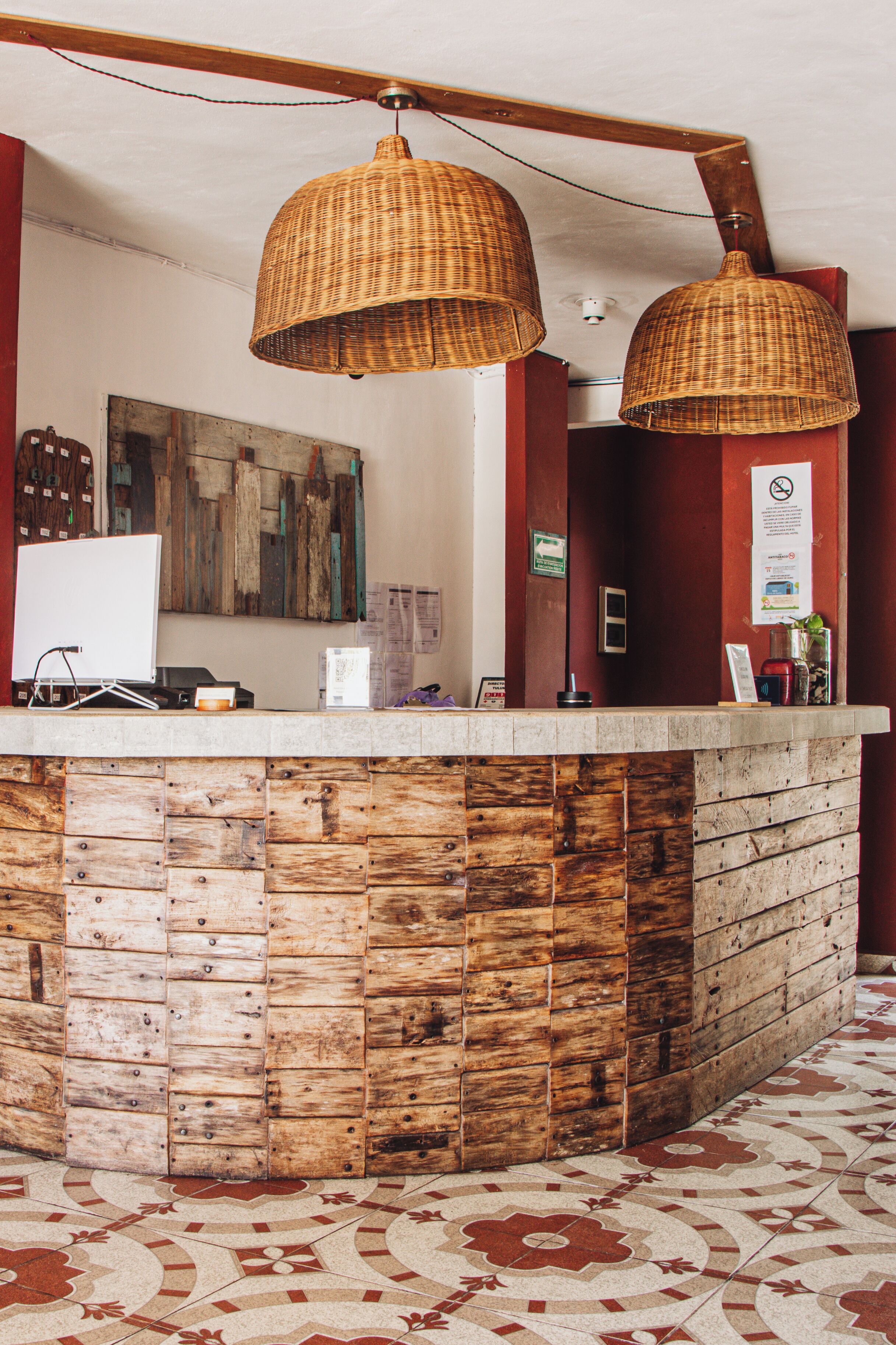 Un comptoir d’accueil au design rustique, en bois, deux suspensions en osier et un sol carrelé orné d’un motif floral.