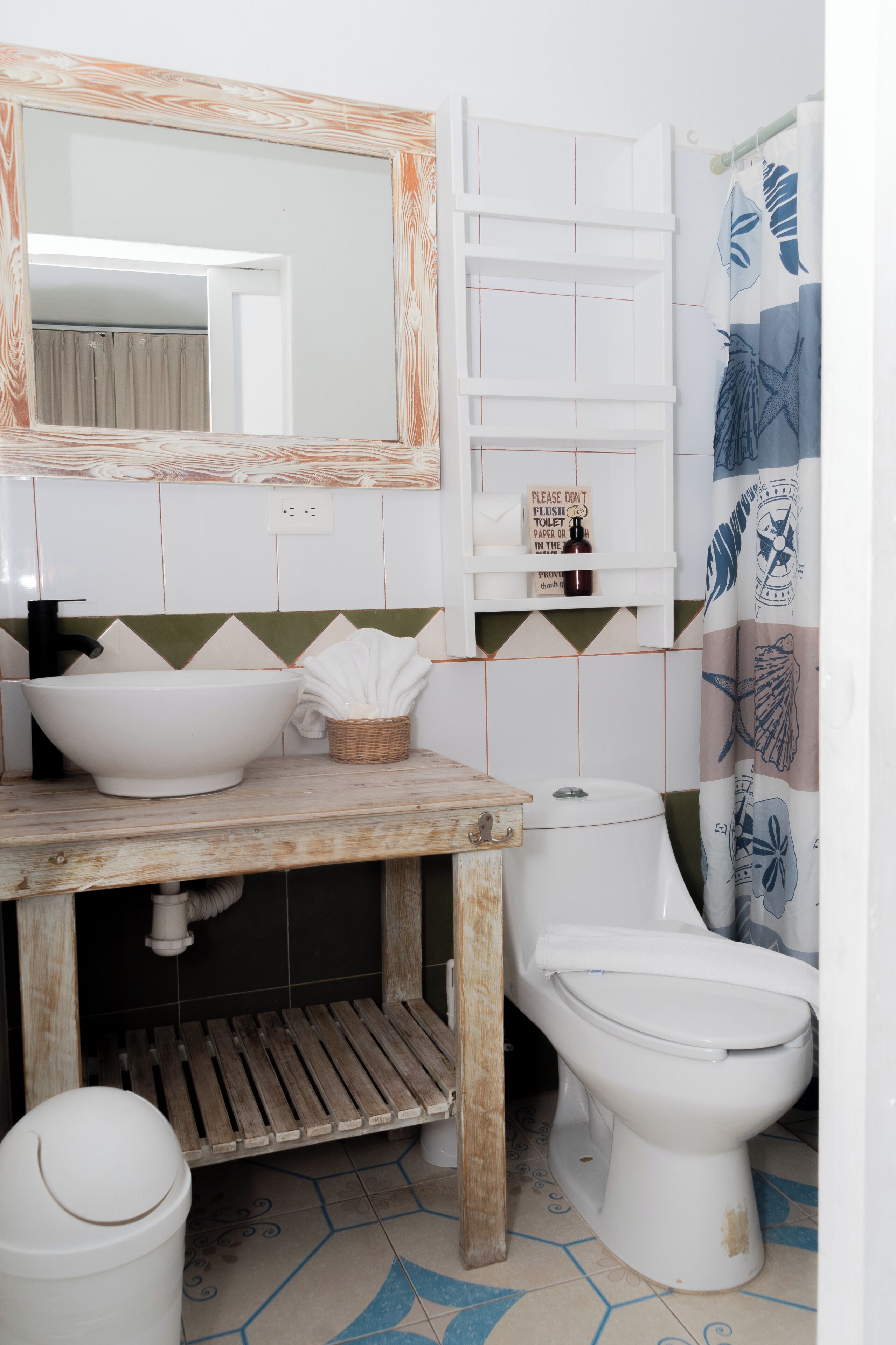 Un baño con tocador de madera, un lavamanos grande de color blanco, inodoro y cortina de ducha con motivos de hojas.