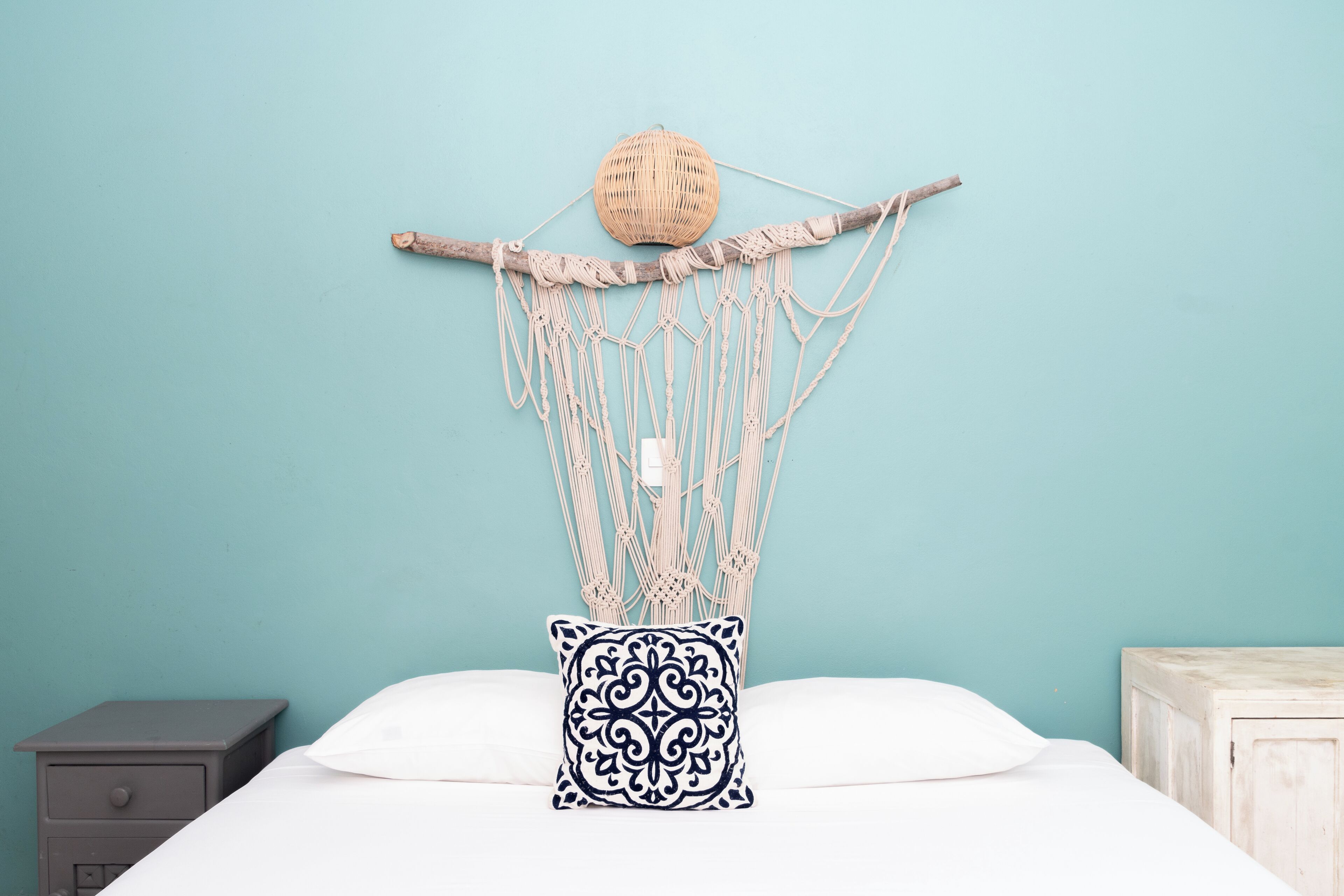A bed with a macrame wall hanging above it, a white pillow with a black pattern, and a small wooden nightstand to the left.
