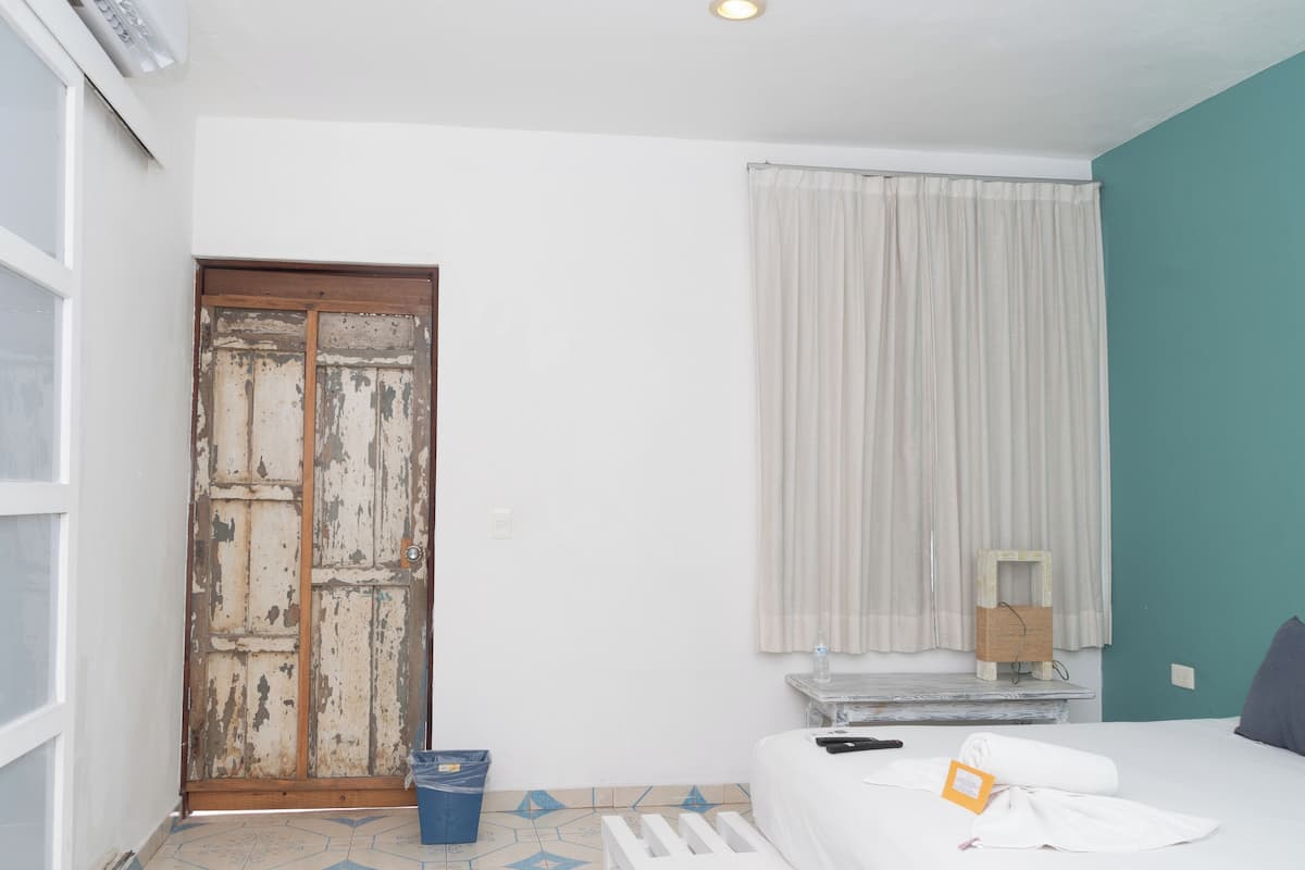 A room with a bed, a window with curtains, a door, a small table with a lamp, and a trash bin.