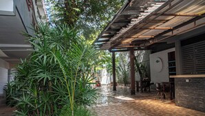 Un espacio al aire libre cubierto con suelo de baldosas, rodeado de vegetación y un edificio de fachada blanca.