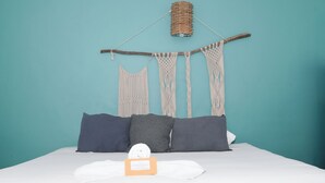 Una cama con sábana blanca, dos almohadas oscuras, un objeto blanco pequeño sobre la cama y un tapiz de macramé en la pared de arriba.